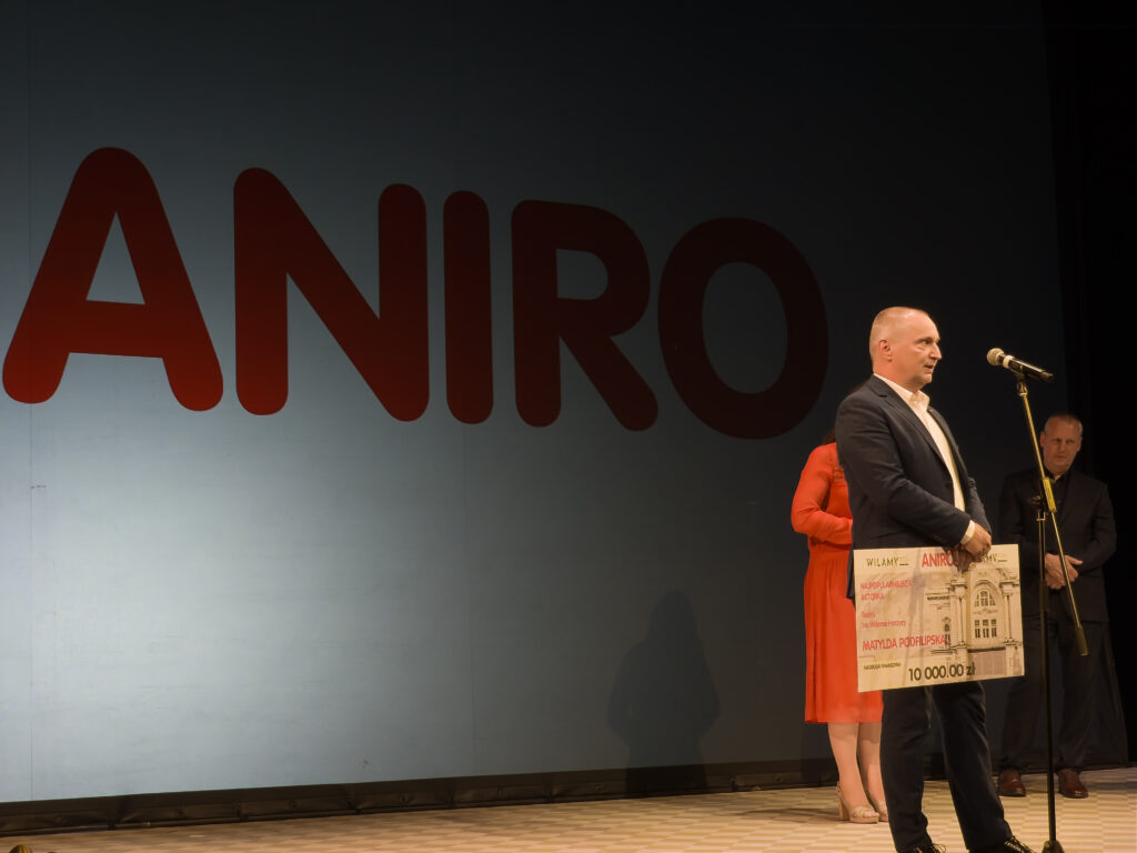 Daniel Kaczmarczyk z ANIRO przemawia przy mikrofonie na Gali Wilamów 2025, prezentując duży czek dla Najpopularniejszej Aktorki. W tle logo ANIRO.